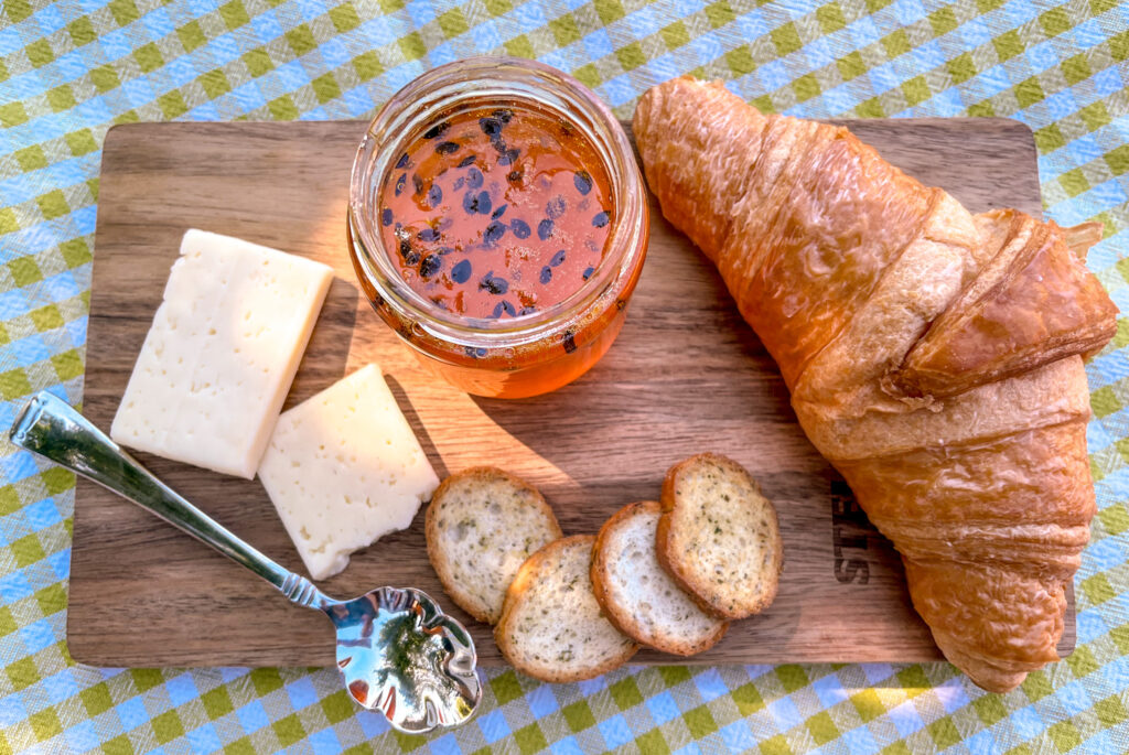 Up-close shot of homemade passion fruit jam on a rustic wooden board with croissant and cheese, on a green and white checkered tablecloth in sunlight with a soft shadow