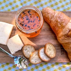 Homemade passion fruit jam on a rustic wooden board with croissant and cheese, on a green and white checkered tablecloth in sunlight with a soft shadow