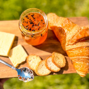 Homemade passion fruit jam on a rustic wooden board with croissant and cheese, held over grass in sunlight