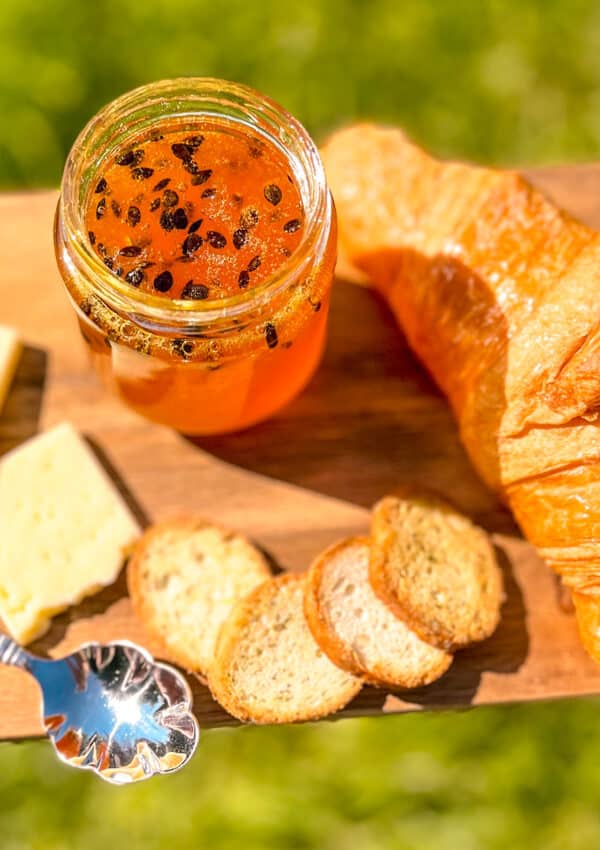 Homemade passion fruit jam on a rustic wooden board with croissant and cheese, held over grass in sunlight