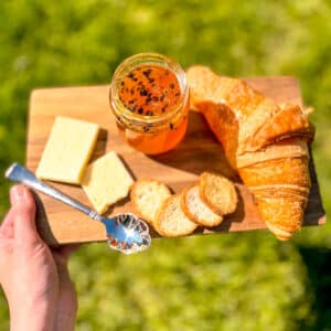 Homemade passion fruit jam on a rustic wooden board with croissant and cheese, held over grass in sunlight