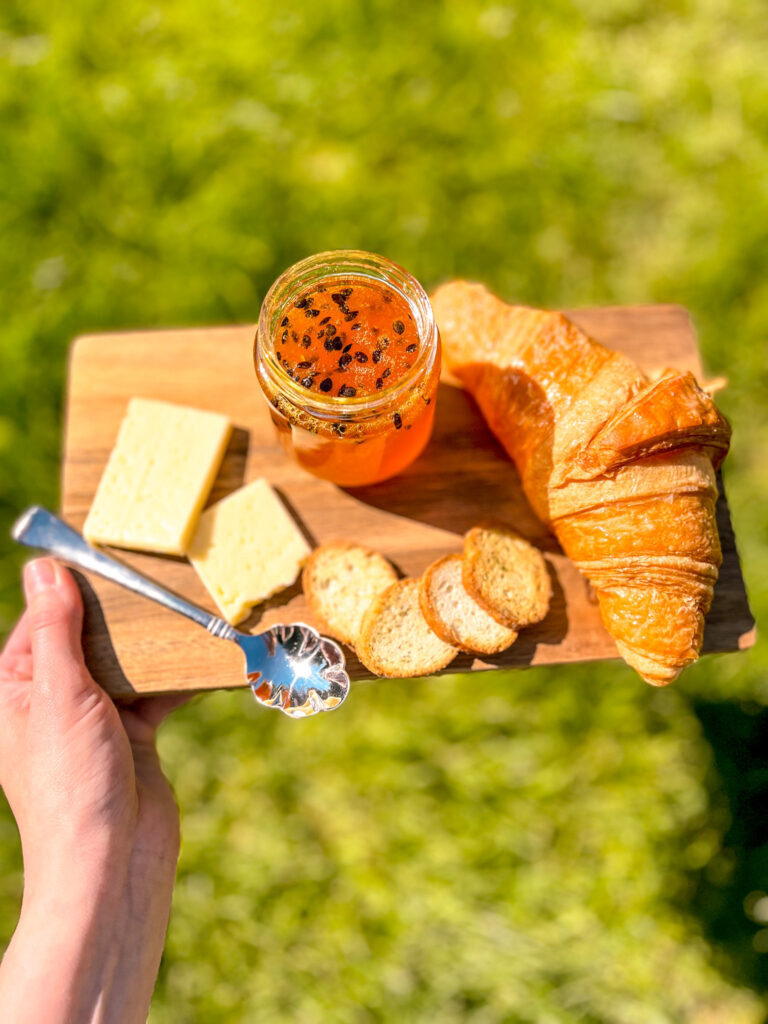 Homemade passion fruit jam on a rustic wooden board with croissant and cheese, on a green and white checkered tablecloth in sunlight with a soft shadow