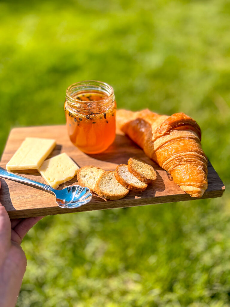 Jar of passion fruit jam on a small wooden cutting board with a croissant and cheese, held outdoors over green grass in bright sunlight.