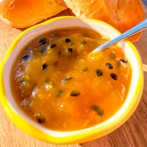 Close-up of homemade mango and passion fruit jam in a jar, with slices of fresh baguette in the background on a rustic wooden surface.
