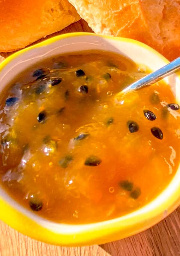 Close-up of homemade mango and passion fruit jam in a jar, with slices of fresh baguette in the background on a rustic wooden surface.