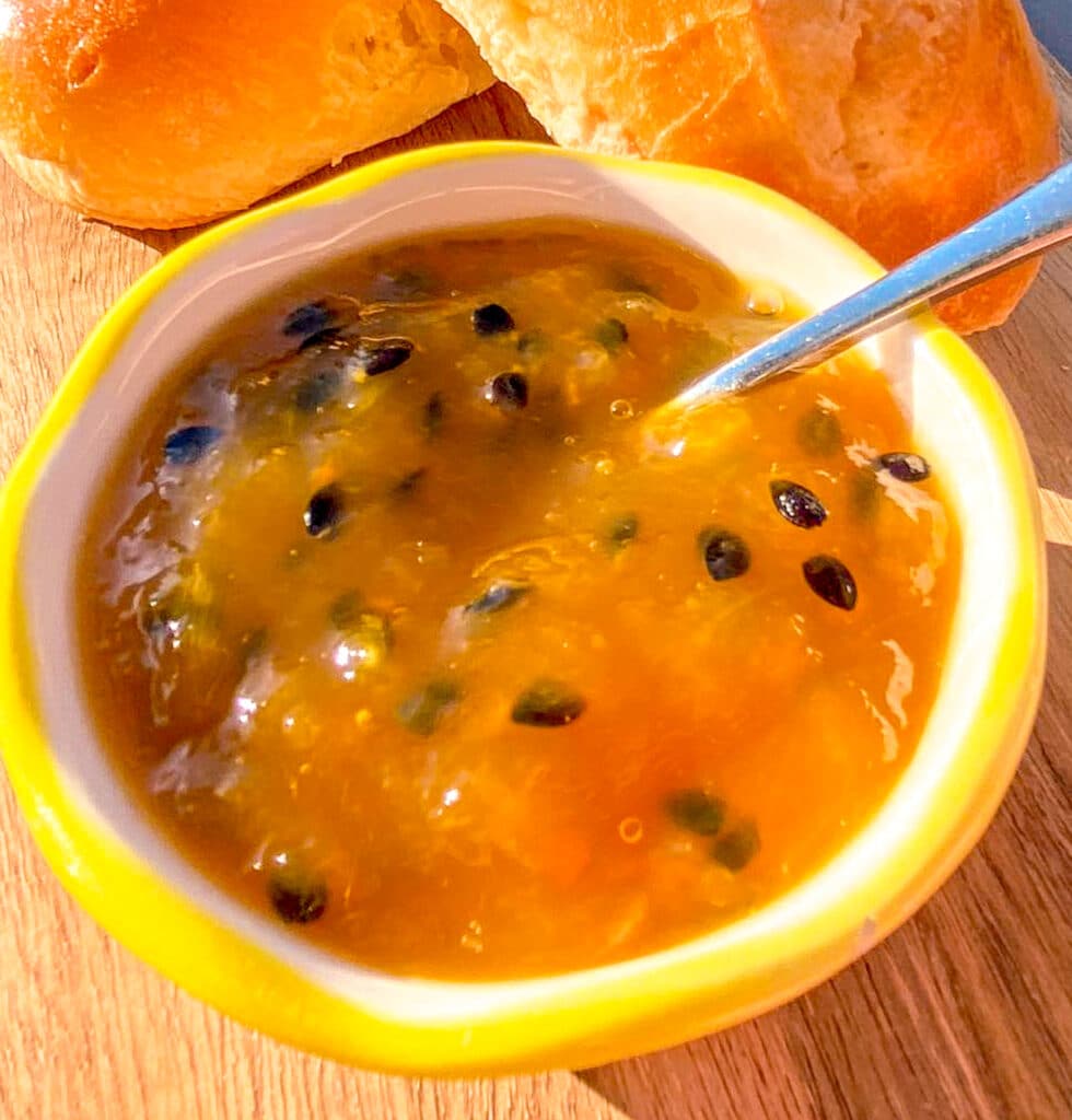 Close-up of homemade mango and passion fruit jam in a jar, with slices of fresh baguette in the background on a rustic wooden surface.