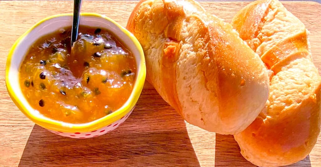  homemade mango and passion fruit jam in a little polka-dot bowl, with slices of fresh baguette in the background on a rustic wooden surface.