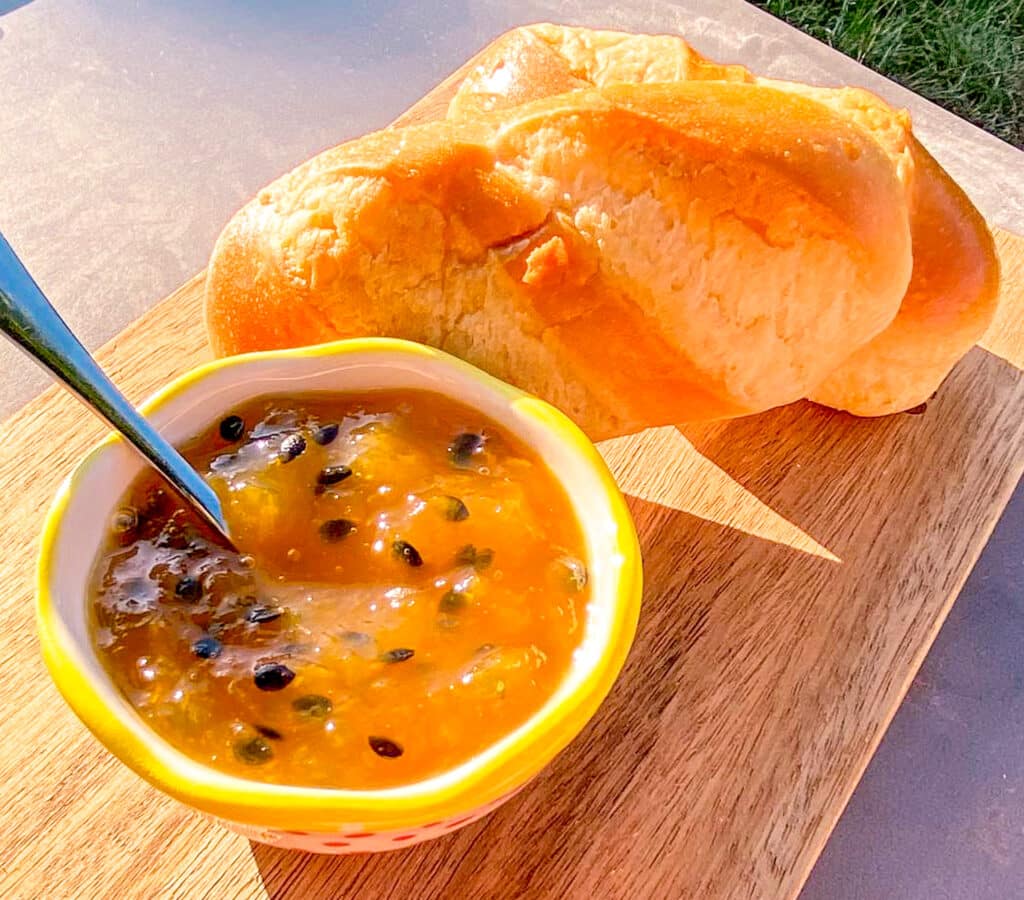  homemade mango and passion fruit jam in a little bowl, with slices of fresh baguette in the background on a rustic wooden surface.