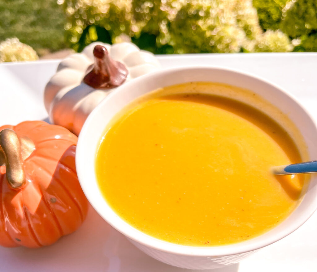 Easy Butternut Squash soup in a white bowl beside two decorative pumpkins near greenery