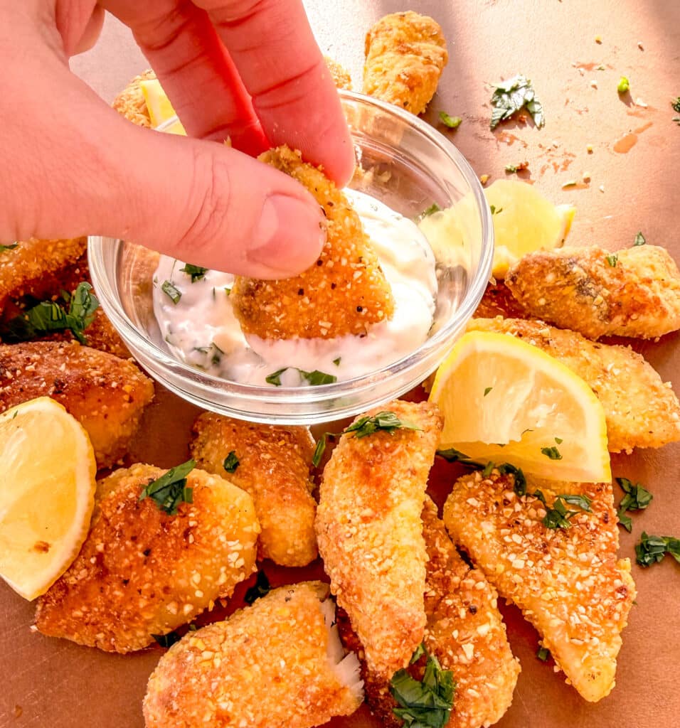 finger dipping a crispy cornmeal tilapia inside a bowl with sauce surrounded by more crispy tilapia and lemons