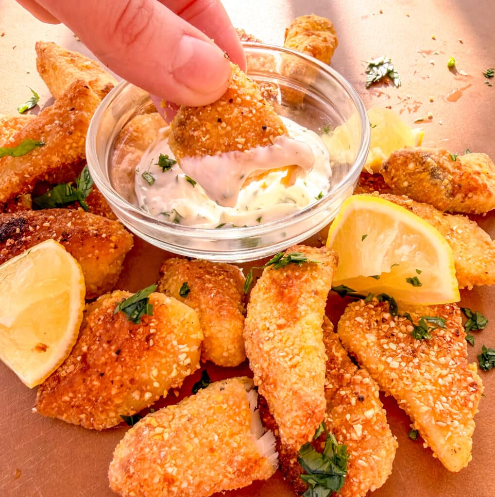 finger dipping a crispy cornmeal tilapia inside a bowl with sauce surrounded by more crispy tilapia and lemons